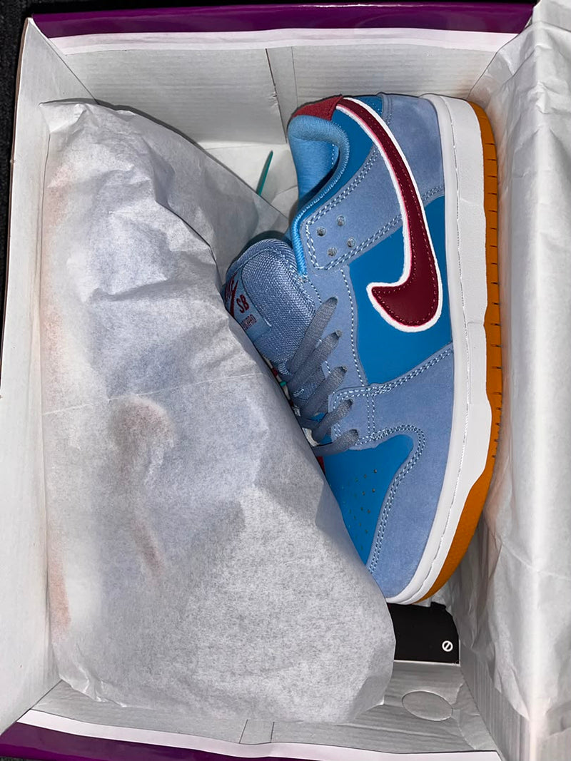 A Nike Dunk sneaker in light blue suede with a burgundy Swoosh, white midsole, and orange gum outsole, partially tucked inside its white shoebox with tissue paper.